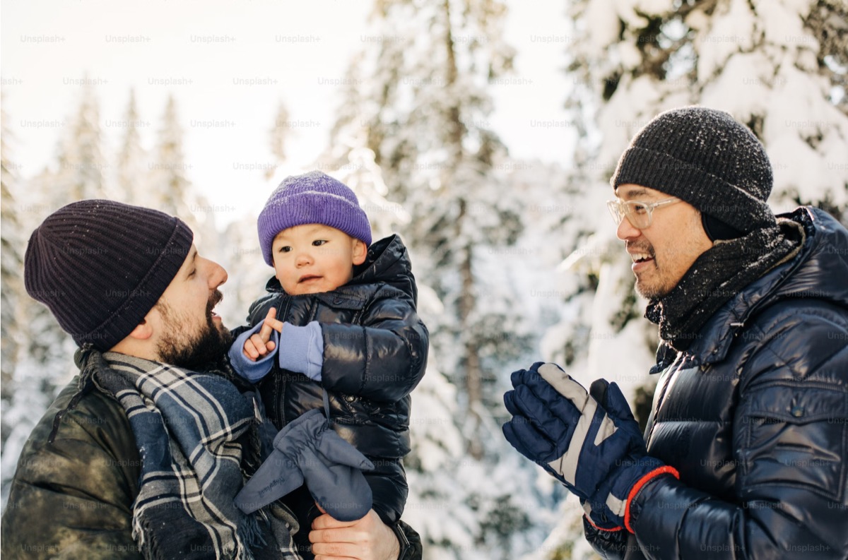 A winter scene featuring three people dressed in warm clothing outdoors with snow-covered trees in the background. One person is holding a young child dressed in a black puffer jacket and a purple beanie, while another person wearing glasses and gloves is engaging with them in conversation.